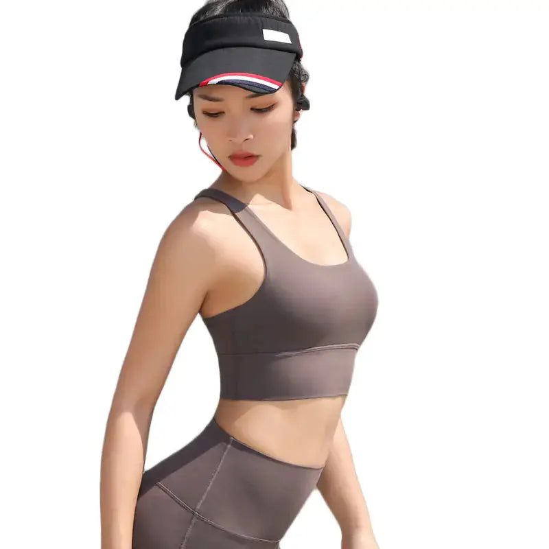 A woman models the My Store Cross-Back Yoga Sports Bra for Beautiful Back Design in taupe, paired with matching leggings and a black visor, as she poses against a white background looking down to her left.