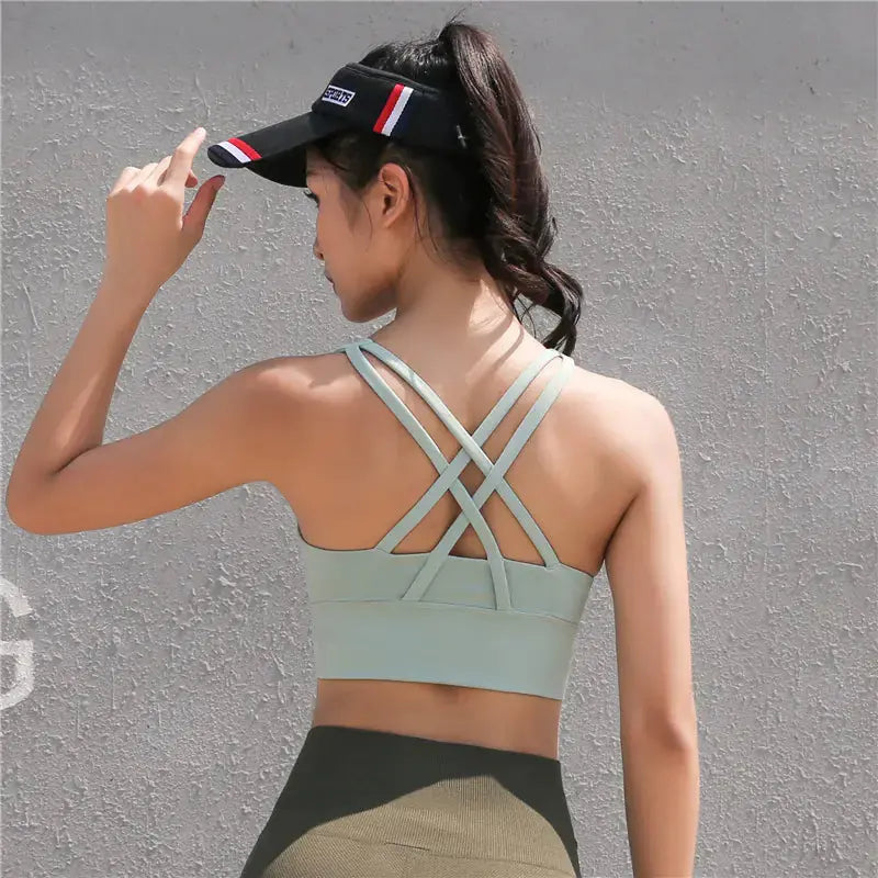 A woman with a ponytail stands against a light gray wall, wearing the My Store Cross-Back Yoga Sports Bra for Beautiful Back Design in light green, paired with olive leggings and holding a black visor.